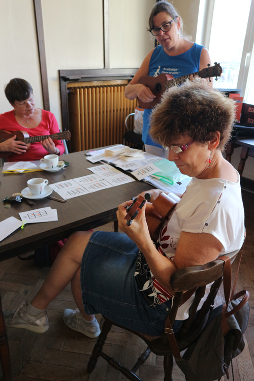 Twee deelneemsters zitten aan tafel met een ukelele in de hand. De lesgeefster staat recht. 