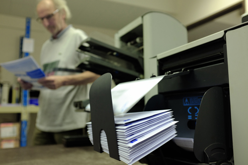 Gros plan d'enveloppes dans une machine. En arrière-plan, quelqu'un regarde une feuille de papier. 